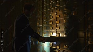 A man is carefully accessing safety deposit boxes in a dimly lit and shadowy vault