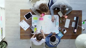 Top view of group of four multiethnic business people sitting at desk in office and using sticky notes to create mind map on large sheet of paper Stock Video