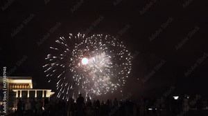 Spectators watch sparkling real fireworks in city square. Pyrotechnic volleys explode in night sky above illuminated building. People attend festival of fireworks and lights in city celebration.