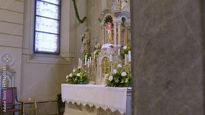 Altar In Church On Wedding Ceremony Day With Golden Candle Holders With Candles on Top of Them, White Roses, Lilies, Orchids, Liturgical Decorations, Religious Sculptures, Stained Glass Windows, Walls