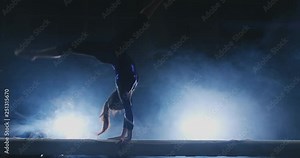 The girl performs a trick on a log in backlight and slow motion in sports gymnastic clothing. Smoke and blue. Jump and spin on the balance beam
