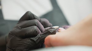 Close up view of nail technician filing client big toe using electric file during pedicure session, with visible dust particles on glove, highlighting attention to detail in foot grooming process | Premium Stock Video Footage