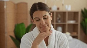 Young woman in spa setting wearing a white robe, looking relaxed and beautiful, indoors at a wellness salon with shelves and plants in the background