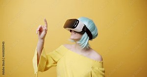 Amazed woman exploring virtual reality. Static shot of astonished young female with blue hair and VR helmet looking around and touching invisible objects against yellow background