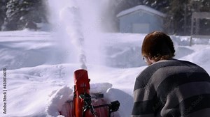 Close up shooting of heaps of fresh snow being removed and shot in the air by a bright orange mechanical snowplough held by a young man in fur hat.