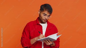 Thoughtful journalist young Indian man making notes, writing down thoughts with pen into notepad notebook diary, to do list, good idea inspiration. Arabian Hindu guy isolated on orange background