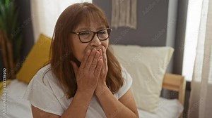 A beautiful mature hispanic woman blows a kiss in her cozy bedroom, smiling with joy while sitting on her bed with pillows in the background.