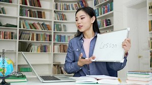 Asian female teacher giving video call lesson using laptop sitting in campus library space. Young woman tutor or coach has remote e-learning for students, online training, a course, talks remotely