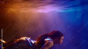 A woman in silver clothing swims underwater in the dark in neon light in slow motion