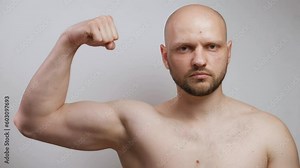 A muscular bald male athlete bodybuilder with a beard and a bare torso looks seriously at the camera, raises his arm, strains and shows his biceps. Sports health, strong physical body