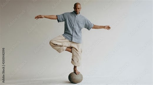 Man balancing on a stone sphere with arms outstretched in a studio setting. A focused individual demonstrates exceptional balance and control while standing on a spherical object