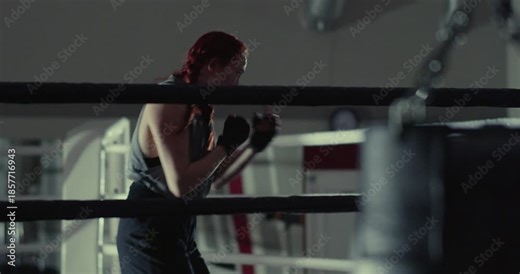 A female boxer's shadow fight drills showcase precision and power beneath dim gym lights. Woman boxer during shadow fighting workout in a dark boxing gym. Cropped slow motion 4k footage.