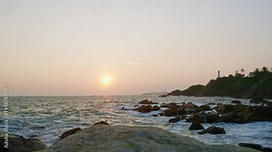 Lighthouse stands on hill at sunset, waves crash against rocks below. Golden hour light reflects on ocean swells, serene nature scene. Coastline video captures tranquil evening by sea. Slow motion.