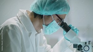 Asian scientist wearing protective mask, sterile hat and gloves using microscope and making notes while conducting research in laboratory