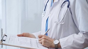 Female doctor taking notes on clipboard. Close-up of a unrecognizable female healthcare professional writing and filling um medication history records form while standing straight in clinic