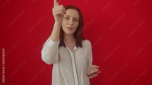 Young and serious caucasian woman pointing at you and camera while standing alone over red isolated background, gesturing with confidence while using smartphone