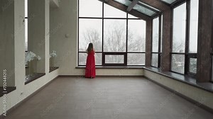 Woman in red dress looks out of winter window. Young female model complements gallery panoramic windows with graceful presence. Charm and elegance