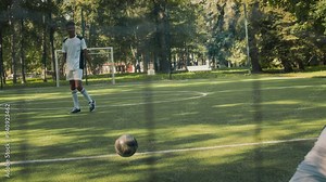 Back view shot of goalkeeper standing by goalpost and punching ball away while playing soccer game on grassy field at daytime