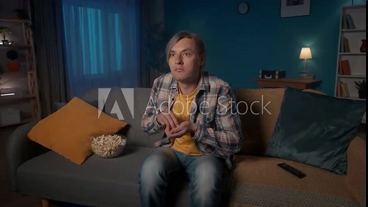 Horror film. Shocked man watching a scary movie on TV at home and eating popcorn. The man shudders in fear and spills the popcorn, screaming in horror, covering his face.