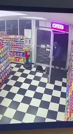 Grainy Security Camera Footage Of Sudden Rottweiler Attack In Local Convenience Store ------------ ----------- ------------- This video presents a tense scene captured through a grainy nineties cctv security camera inside a quiet convenience store at night. The interior features a classic black-and-white checkered floor and shelves stocked with colorful snacks, all illuminated by harsh fluorescent overheads and a faint pink neon glow. A man dressed in a dark blue hoodie and black pants enters th