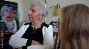 Elderly woman looks off camera and talks to a mature woman with eyes rolling.