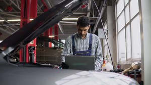 Experienced indian mechanic in uniform is using a laptop while repairing car in auto service