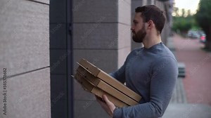 Hand held shot of delivery man coming to door and rings doorbell. Bearded courier holding cardboard boxes with pizza. Courier provides fast delivery of hot food to client. Shooting in slow motion.