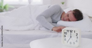 Sleeping man lying on bed in his bedroom and stopping alarm clock at home. Wake up in morning of working day. Young sleepy guy reaching for alarm clock to switch it off. 4k video. Slow motion video.