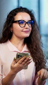 Attractive lady in a good mood has correspondence with somebody on the phone. Woman is thinking for a while what to write. Office staff at desk at the backdrop in blur. Vertical video