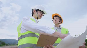 A senior manager in eyeglasses teaching a young attractive trainee on an unfold blueprint, labor and employment, diversity people in workplace, engineering training, safety uniform, sustainable energy
