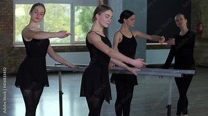 Young woman dancers in black tights practicing graceful movements of classical choreography while standing at ballet barre. Diligent ballerinas training with choreography teacher in dance hall
