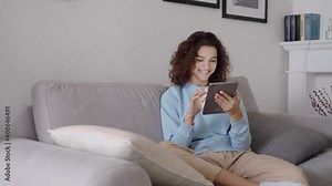 Happy hispanic teen girl holding pad computer gadget using digital tablet technology sitting on couch at home. Smiling young woman using apps, shopping online, reading news, browsing internet on sofa.