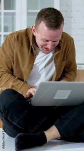 Young man looks mad working long hours online. Guy sitting on a kitchen table and typing on a wireless laptop. Funny fellow playing computer games at home. Vertical video Stock Video