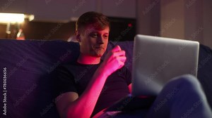 Hipster male inhaling e-cigarette vaping device sitting on couch looking to laptop computer screen in dark living room. Relaxed handsome man smoking electronic cigarette vapor, enjoy moment at home.