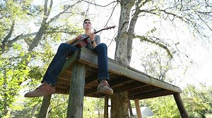 Teenage boy plays guitar on a tree platform, backlit by bright afternoon sun.