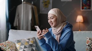 Smiling modern young muslim woman in hijab scrolls social networks on smartphone. Happy islamic arab covered girl young-adult scrolling social media smartphone, housewife leisure, outrage disapproval