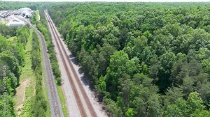 Daytime overhead view from a drone of two parallel railroad train tracks and a commuter passenger train near fields and industry in a rural location.