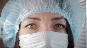 Tired look of a health worker wearing a surgical mask during a break. Overworked healthcare professional. young female doctor in protective medical mask on her face and cap on her head. Close up.
