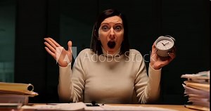 Woman works late at night with a stack of papers and an alarm clock