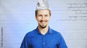 Person with tin-foil hat smiling into camera 4K. Static portrait shot of a man in focus wearing a blue elegant shirt and a tin-foil hat, smiling into the camera.