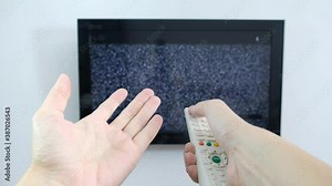 Hand with TV remote control in front of the screen with white noise on it - tuning the television channels and connecting problems