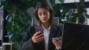 Young businesswoman holds contemporary smartphone and starts entering digits using business card. Concentrated office worker takes moment to make call using phone number provided on bank card
