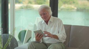 Smiling senior elder 80s man using digital tablet computer pad modern tech relax on sofa. Happy retired old grandfather browse internet read e book in app chatting online with distance doctor at home