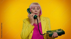 Senior stylish granny woman talking on wired vintage telephone of 80s, says hey you call me back. Elderly old grandmother in yellow jacket isolated on orange background. People lifestyle emotions
