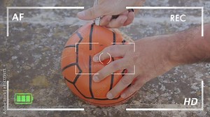 Seen through the viewfinder of a recording camera: piercing a basketball with a pair of sharp scissors, deflating the toy. Exterior day shot.