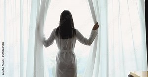 Back rear view at happy young woman wear night robe come to window open lace curtains looking outside dreaming feel motivated rich healthy in modern home hotel bedroom start morning lifestyle concept