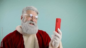 Modern Santa Claus. Cheerful Santa Claus is talking on the phone, by video call, at a distance, he says hello, wishes you merry christmas. blowing snow into telephone. blue background. slow motion
