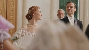 Medium shot of diverse pairs of beautiful debutantes and their male partners wearing adorable Regency outfits strolling along luxury ballroom before dancing