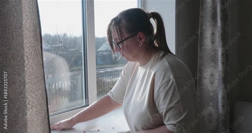 A focused woman wearing glasses carefully wipes a white windowsill with a damp cloth while doing housework. The woman removes dirt and dust from the windowsill to keep the house clean.