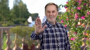Smiling old man shows his palm as a sign of rejection. Summer garden in the background.